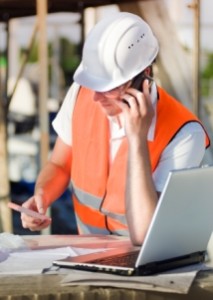 Trade construction worker in hi-vis jacket, helmet, on mobile phone using laptop