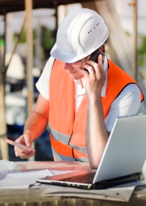 Trade construction worker in hi-vis jacket, helmet, on mobile phone using laptop